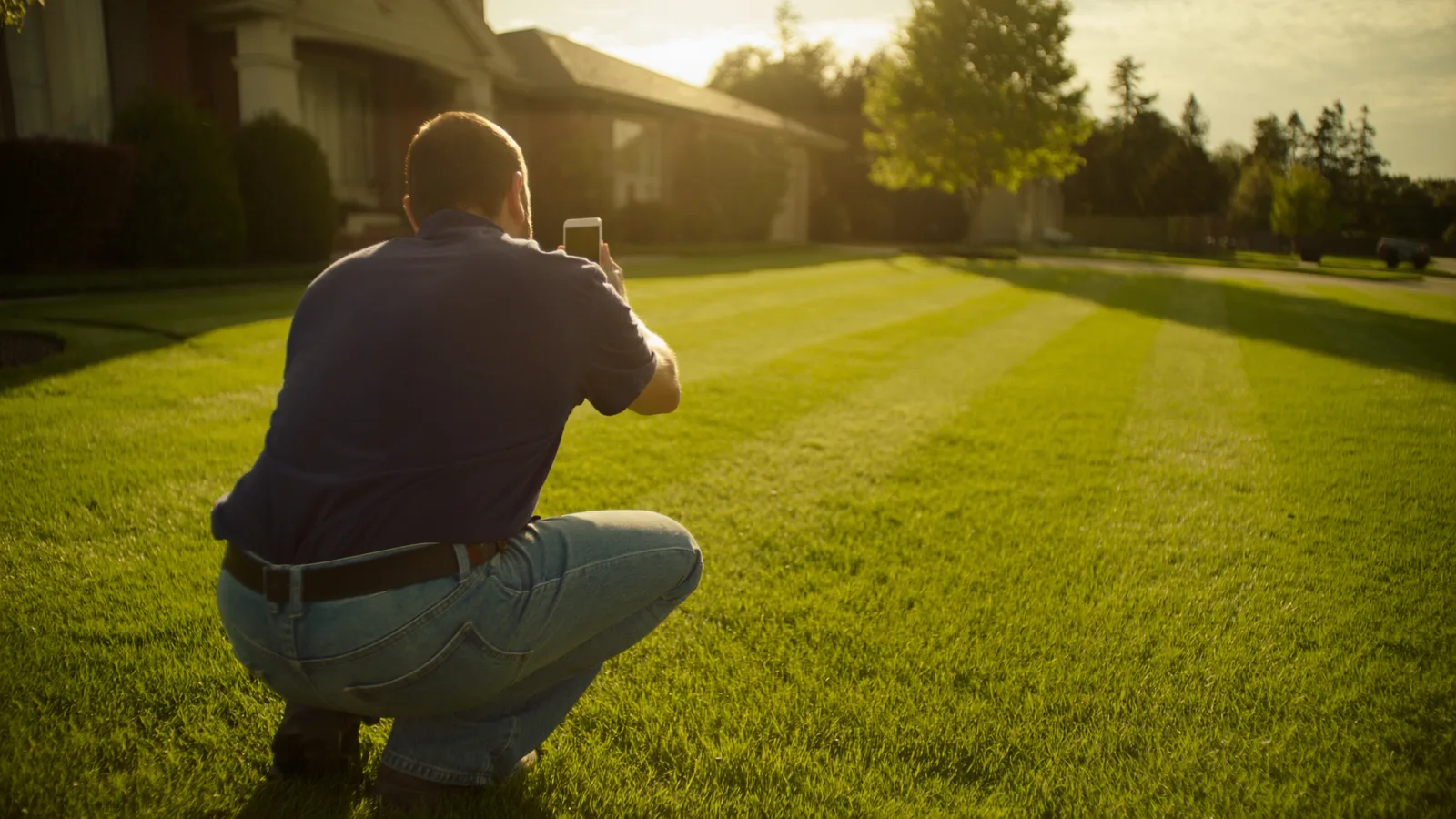 A lawn care professional crouching low at golden hour to photograph his freshly striped work in front of a suburban home